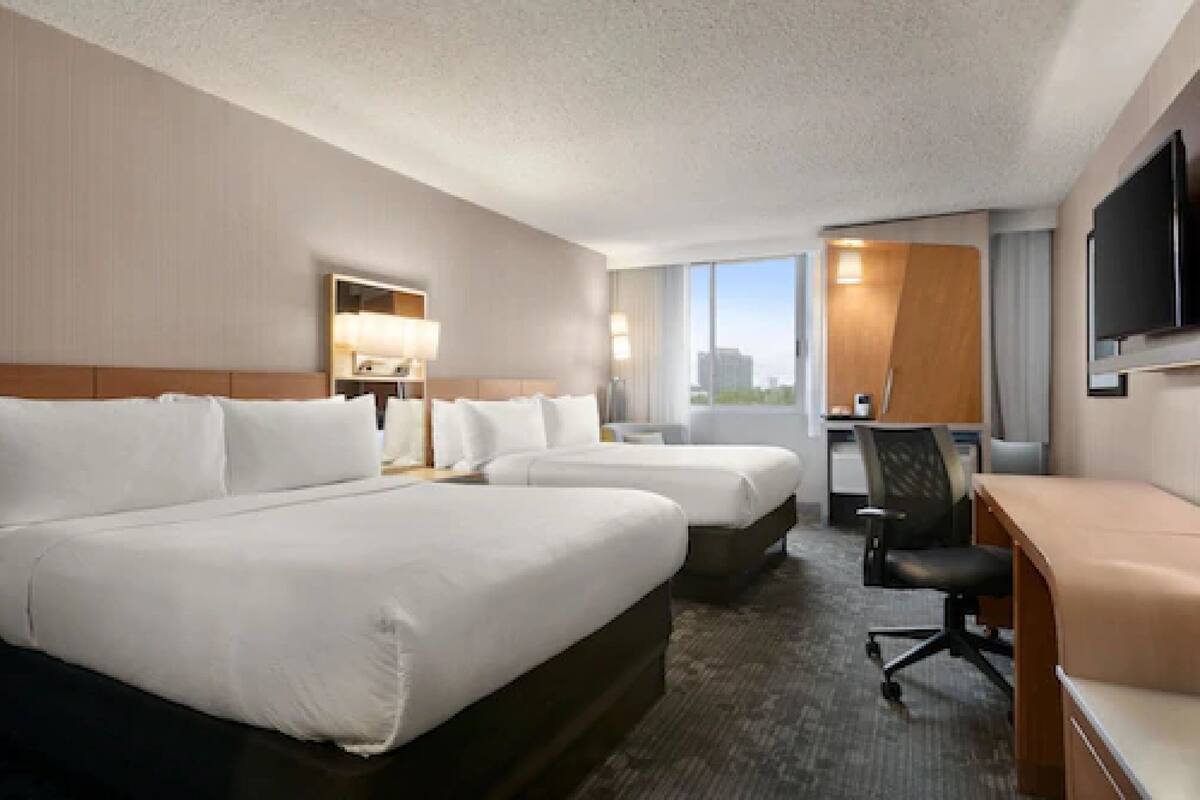 A hotel room with two beds, a desk, a chair, a TV, and a large window with a city view.