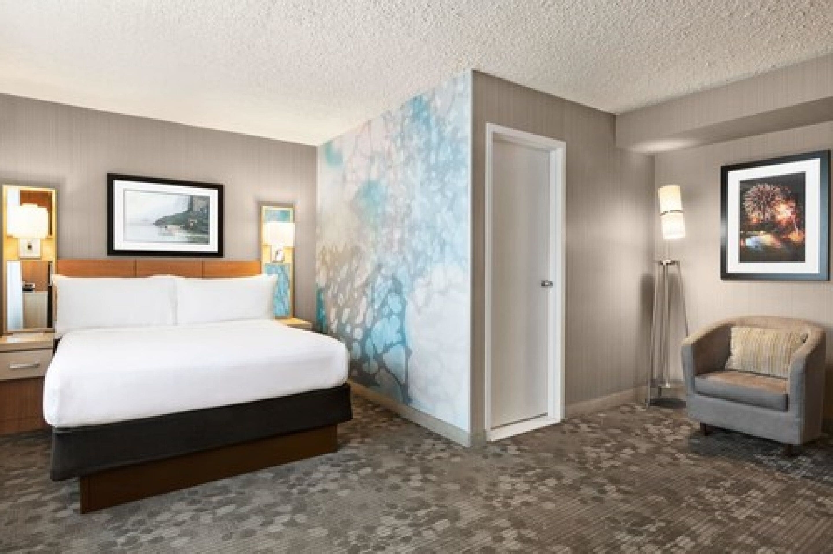 A hotel room with a large bed, a nightstand, a chair, and a wall with a blue abstract design.