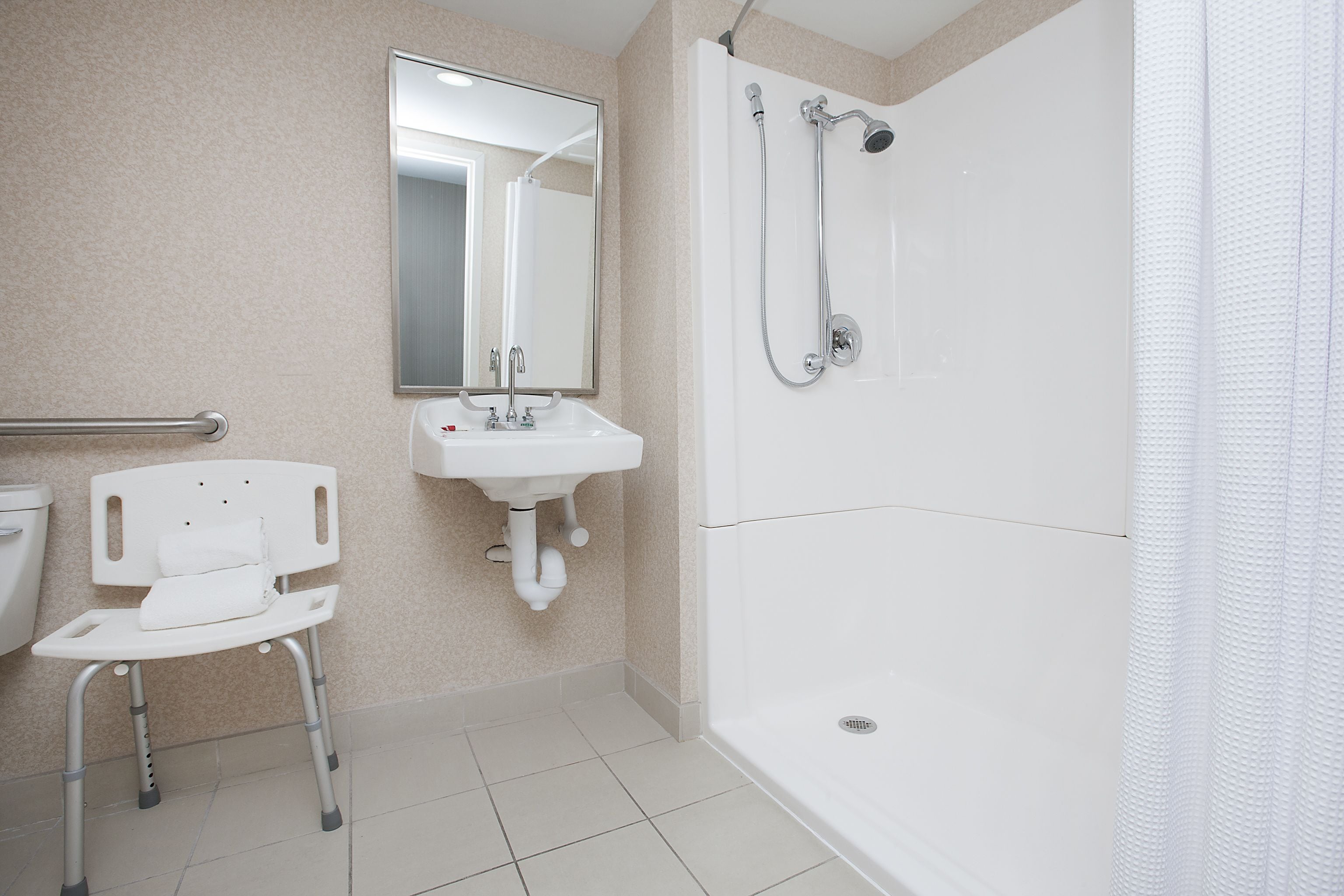 A bathroom with a white sink, mirror, and shower. There is a wheelchair in the corner.