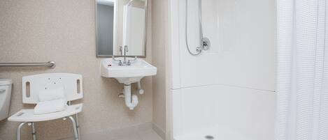 A bathroom with a white sink, mirror, and shower. There is a wheelchair in the corner.