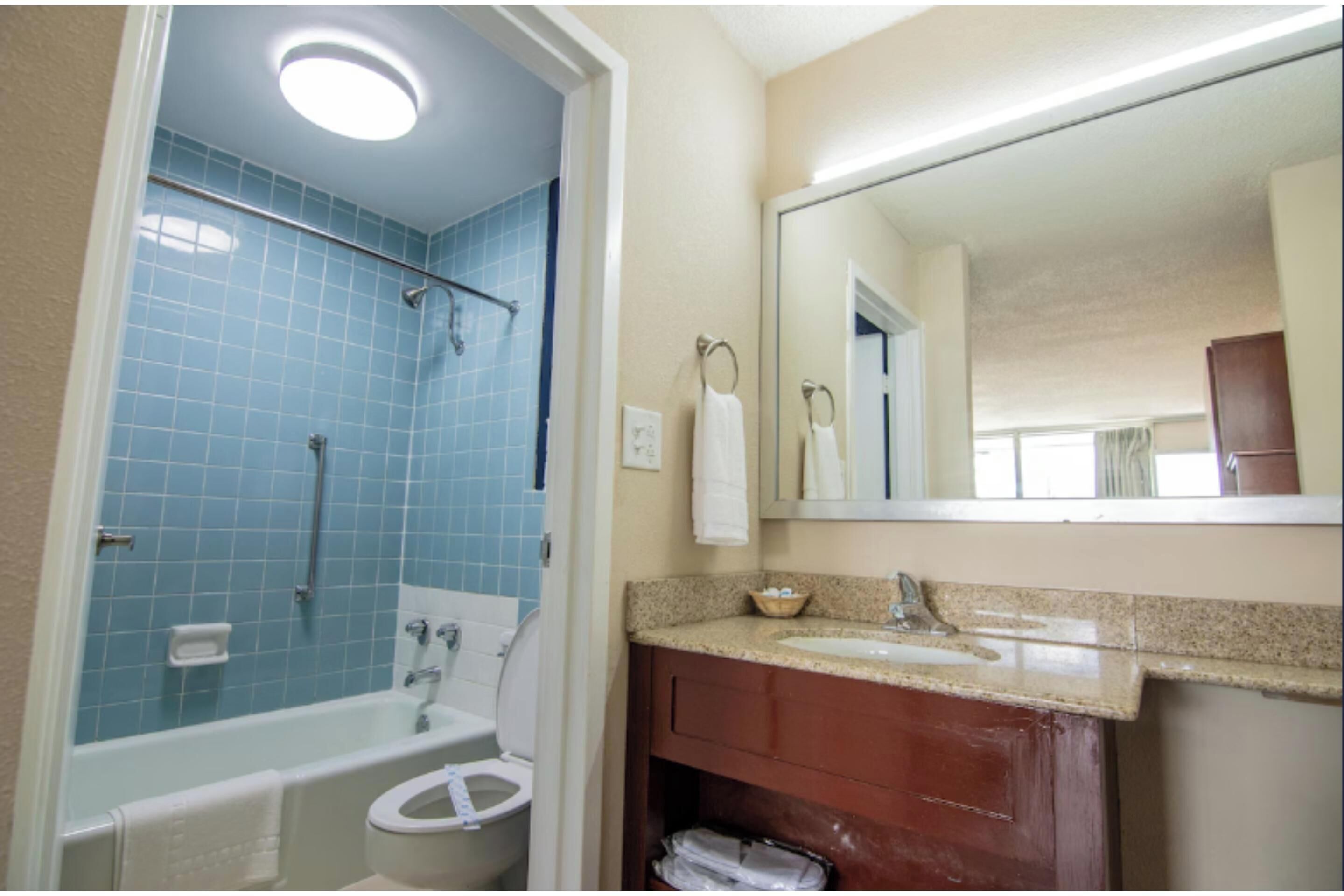 Un baño con ducha de azulejos azules, inodoro blanco, lavabo con tocador de madera y espejo sobre el tocador.