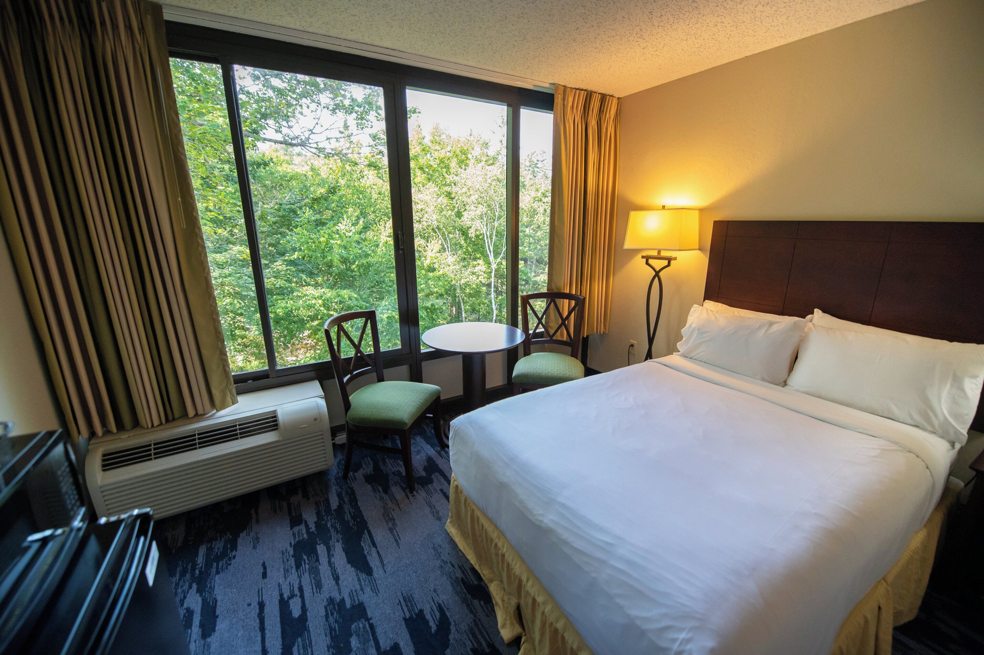 Une chambre d’hôtel avec un lit, une table, deux chaises, une lampe et une fenêtre avec des rideaux.