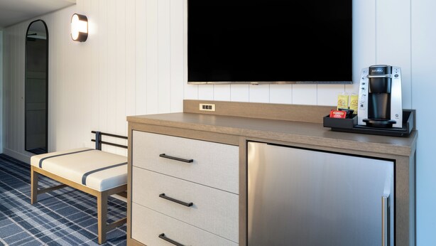 Ein modernes Hotelzimmer mit Flachbildfernseher an der Wand, einer Minibar, einer kleinen Sitzecke und einer Kaffeemaschine.
