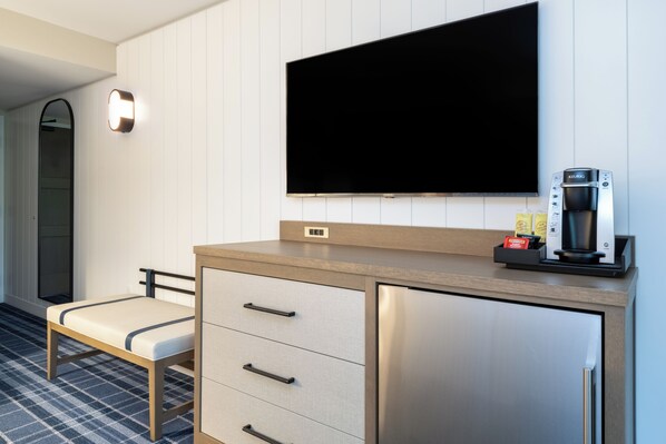 Ein modernes Hotelzimmer mit Flachbildfernseher an der Wand, einer Minibar, einer kleinen Sitzecke und einer Kaffeemaschine.