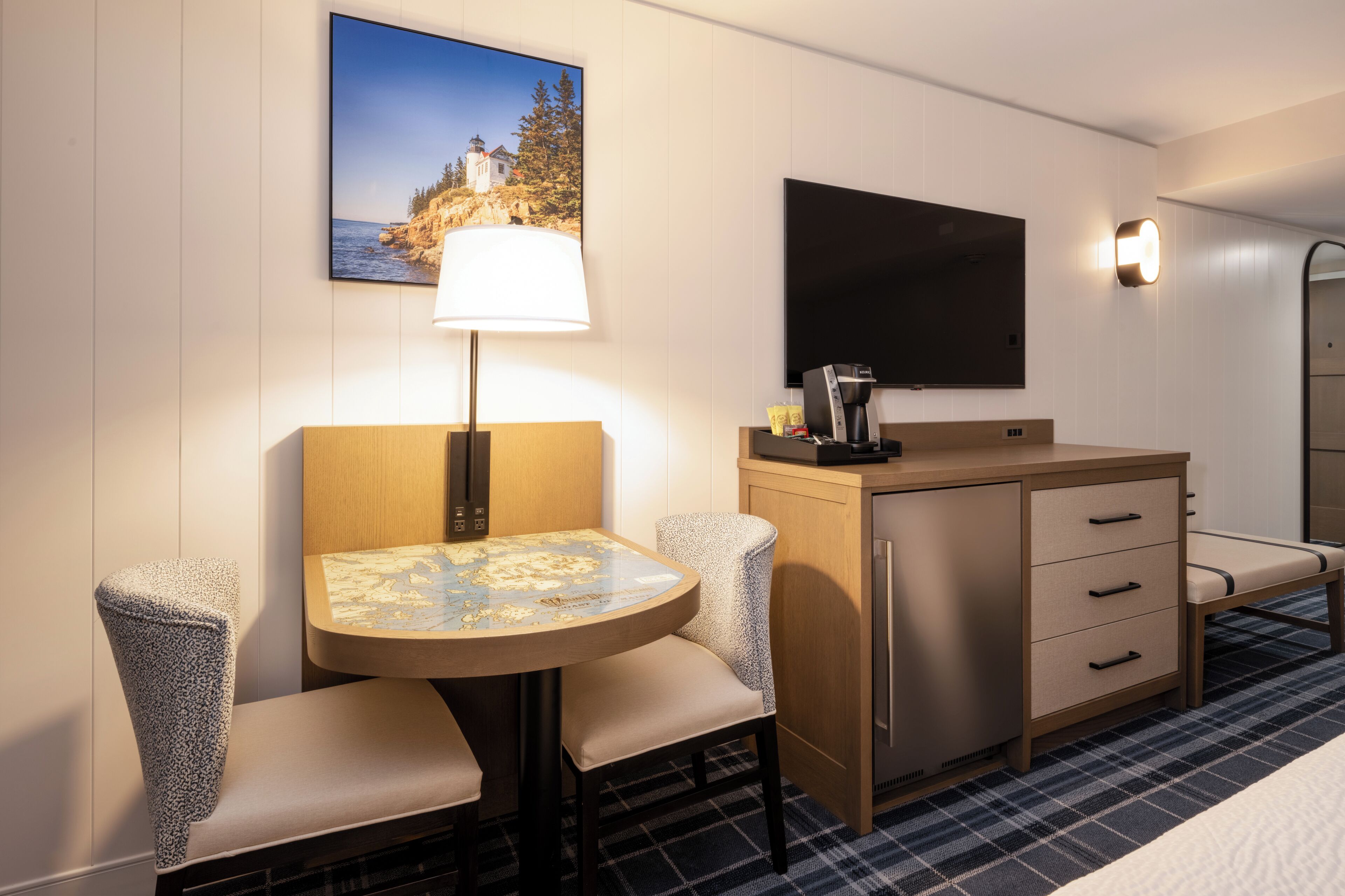 A hotel room with a round table, two upholstered chairs, a small cabinet, a flat-screen TV, and a painting of a coastal landscape.