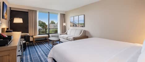 A hotel room with a large bed, a desk with a lamp, a chair, a small table, and a view of the outdoors.