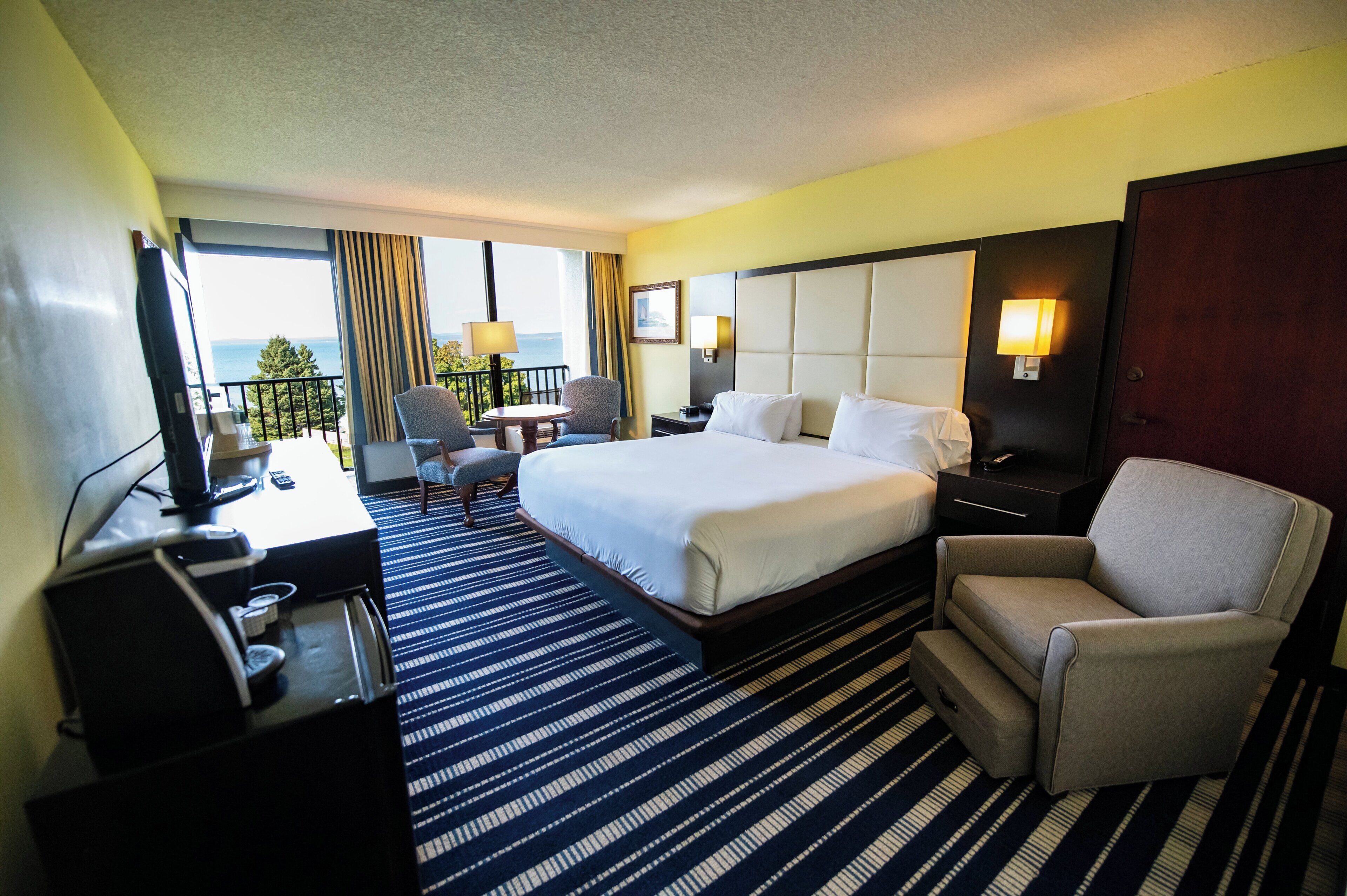 A hotel room with a large bed, a desk with a chair, a small table, and a balcony with a view.