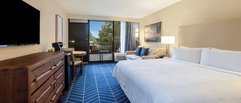 A hotel room with a large bed, a dresser, a desk, a chair, a TV, and a balcony with a view of trees.