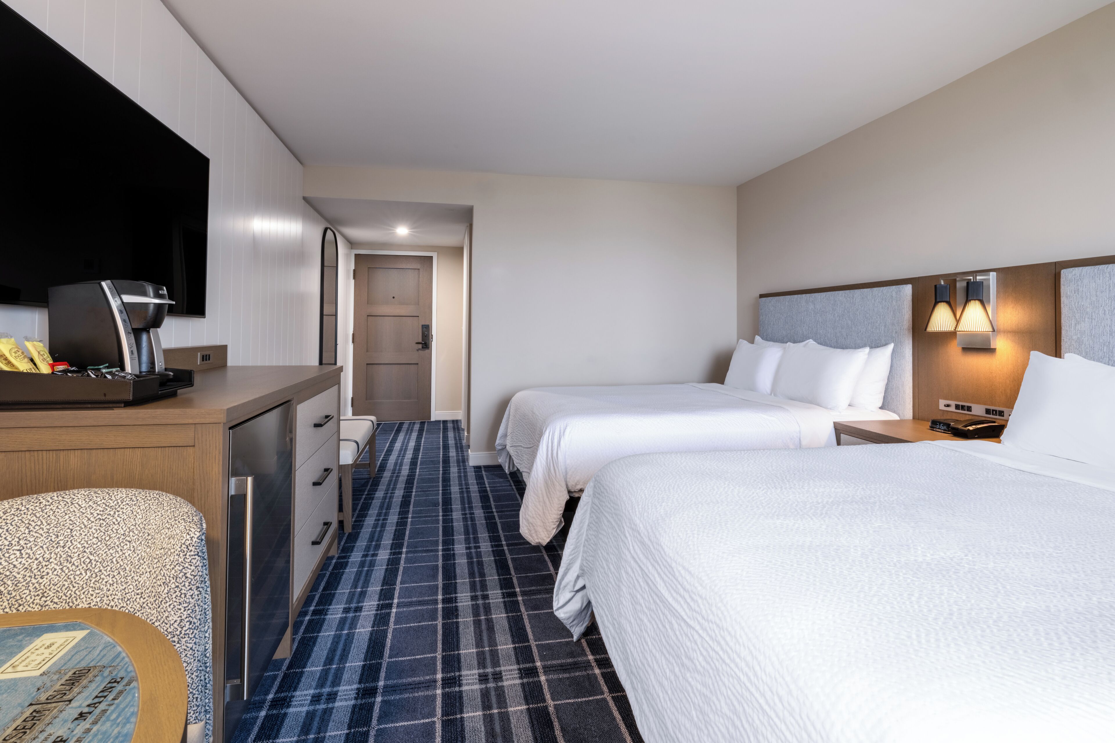 A hotel room with two beds, a desk with a coffee maker, a TV, and a small table.