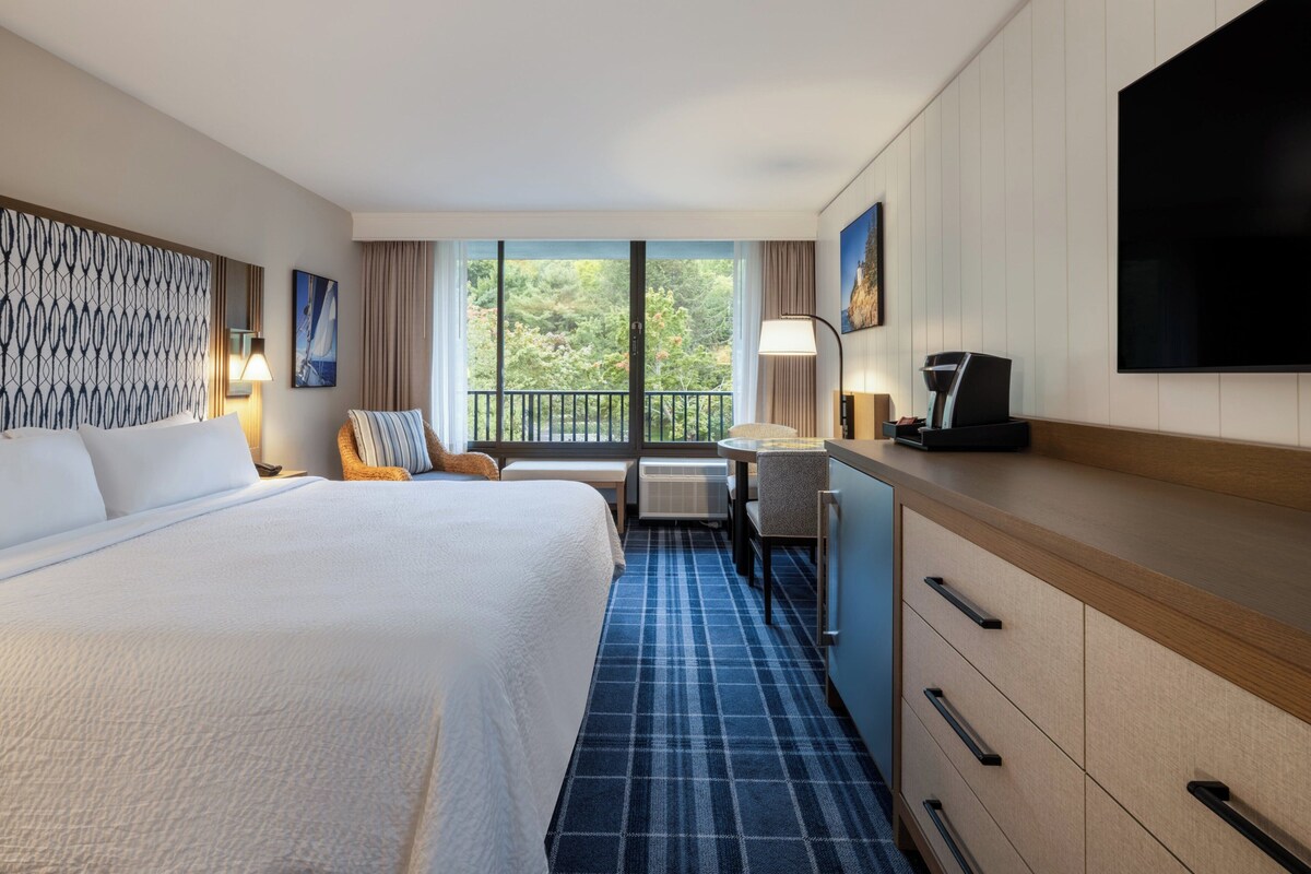 Ein Hotelzimmer mit einem großen Bett, einem Schreibtisch mit Stuhl, einem Flachbildfernseher und Blick auf Grün durch große Fenster.