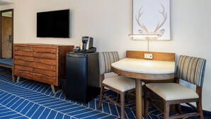 Ein Hotelzimmer mit einem Holzkasten, einem kleinen schwarzen Kühlschrank, einem Flachbildfernseher, einer Dekoration aus einem gerahmten Hirschgeweih, einem runden Tisch mit zwei Stühlen und einem blau gemusterten Teppich.