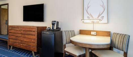 A hotel room with a wooden dresser, a small black refrigerator, a flat-screen TV, a framed deer antler artwork, a round table with two chairs, and a blue patterned carpet.