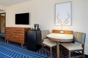 Ein Hotelzimmer mit einem Holzkasten, einem kleinen schwarzen Kühlschrank, einem Flachbildfernseher, einer Dekoration aus einem gerahmten Hirschgeweih, einem runden Tisch mit zwei Stühlen und einem blau gemusterten Teppich.