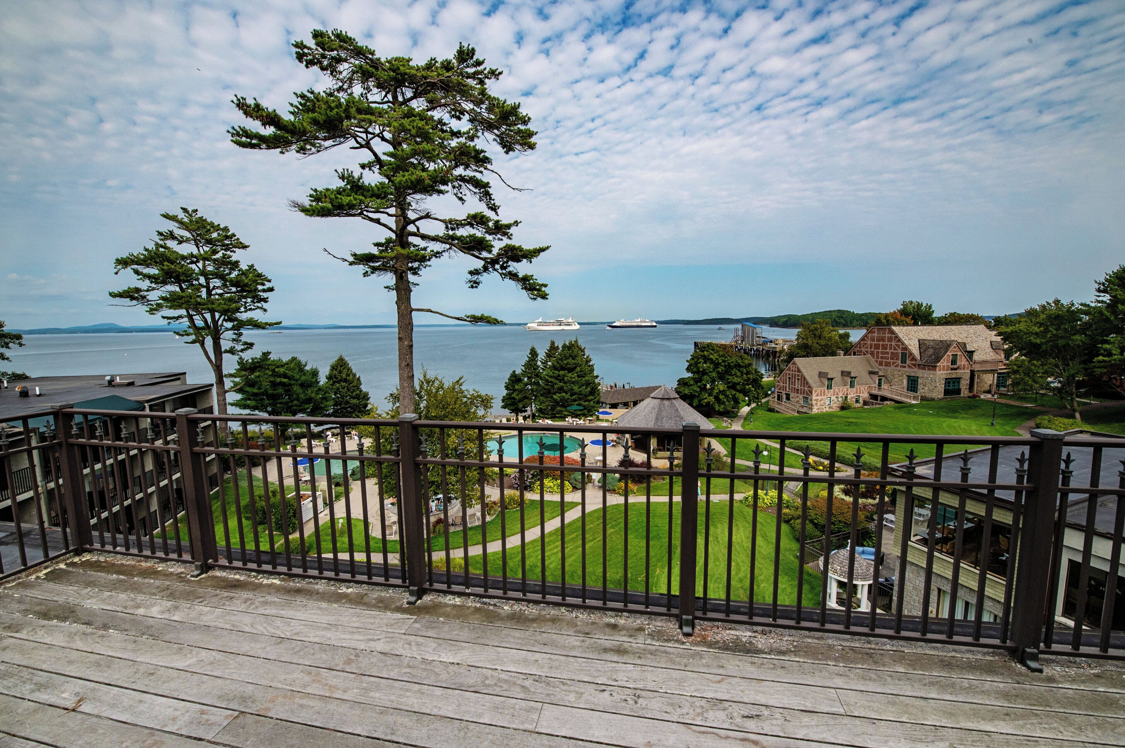 A view from a balcony with a wooden deck, overlooking a pool area, a large body of water, and a distant ship.