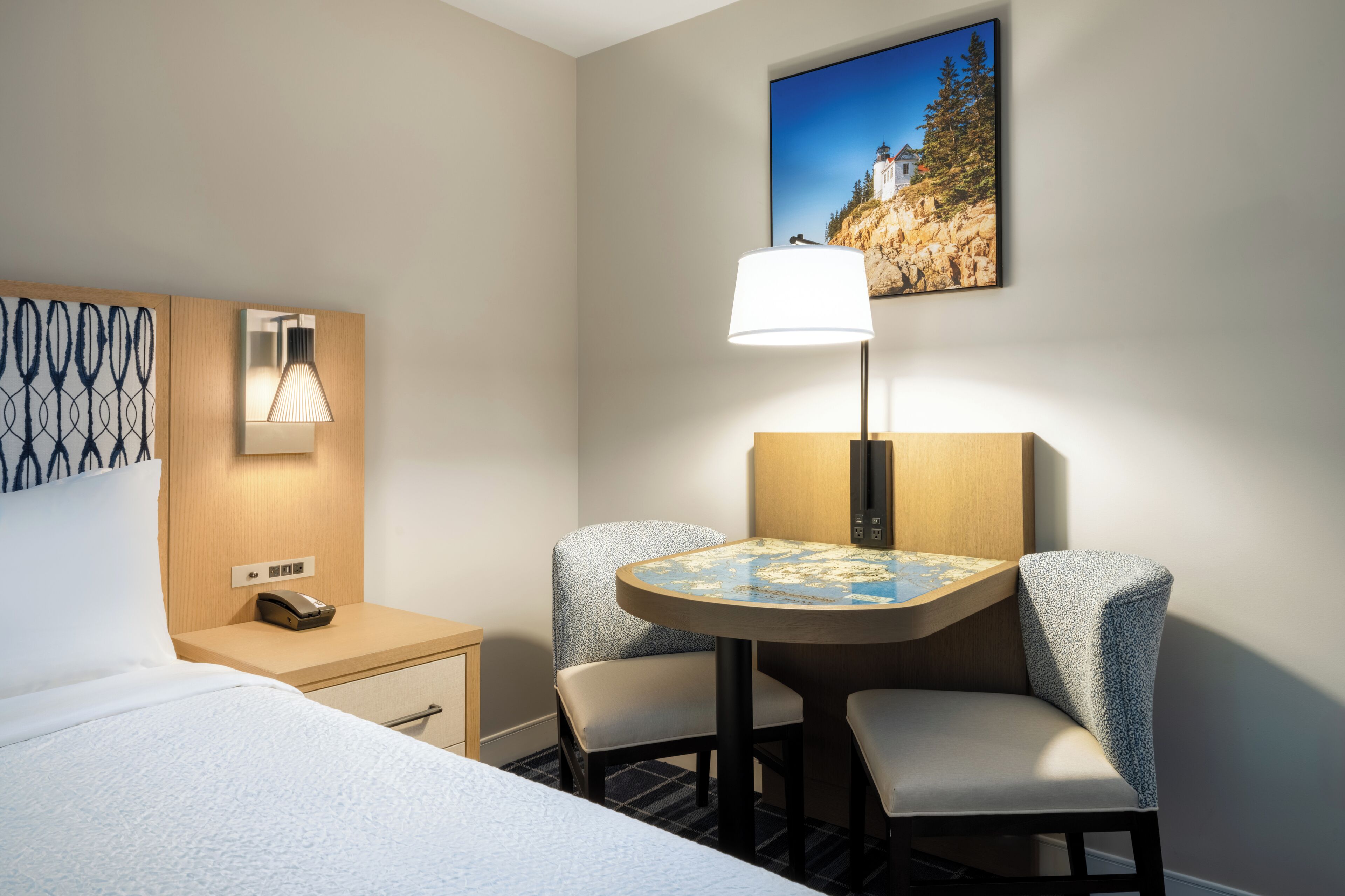 A hotel room with a bed, a desk with a lamp, two chairs, and a painting on the wall.