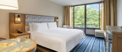 Une chambre d’hôtel avec un lit, une table ronde, une chaise, un bureau et une grande fenêtre avec des rideaux.