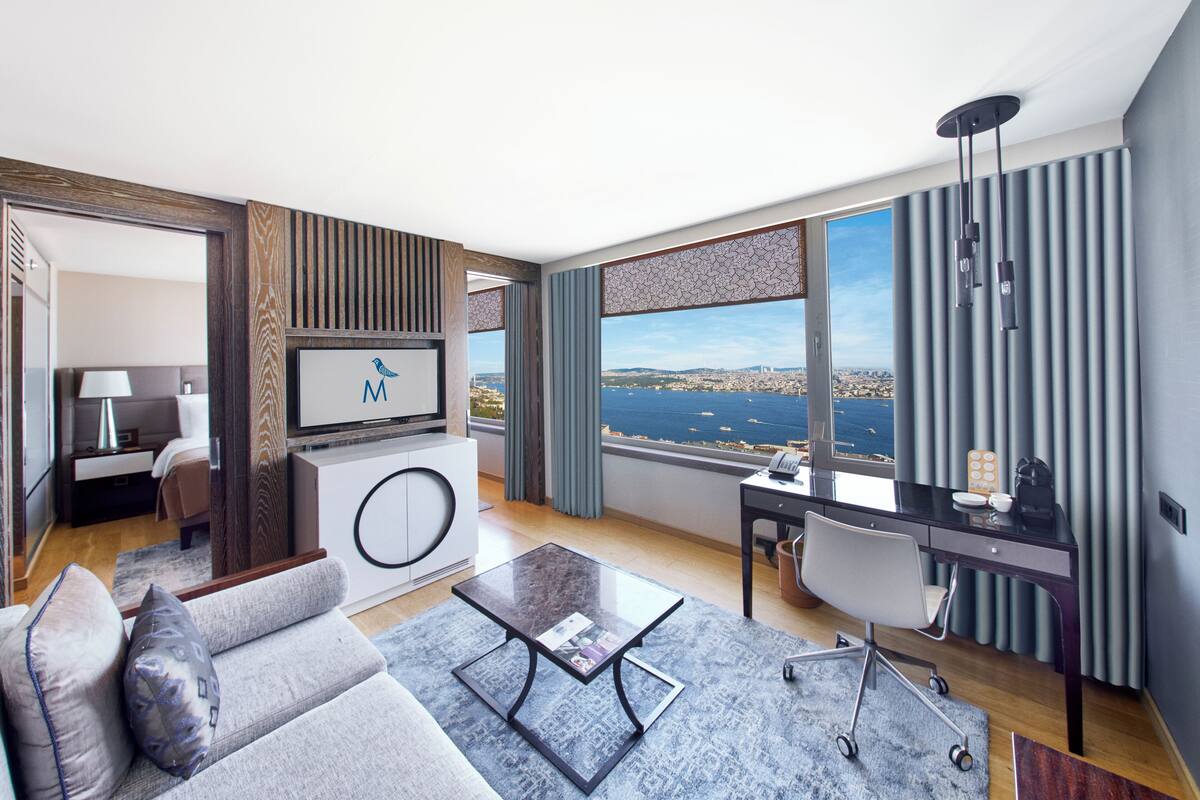 A modern hotel room with a large window offering a city view, a comfortable sofa, a desk, and a television.
