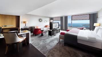 A hotel room with a bed, a desk with a chair, a television, and a view of the sea.