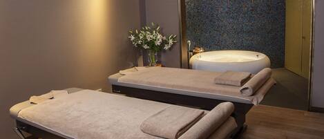 A spa room with two massage tables, a large bathtub, and a mirror reflecting the room.
