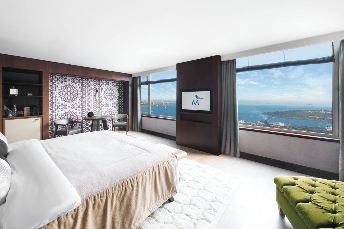 A modern hotel room with a large bed, a green ottoman, a desk with a chair, and a view of the city and sea through large windows.