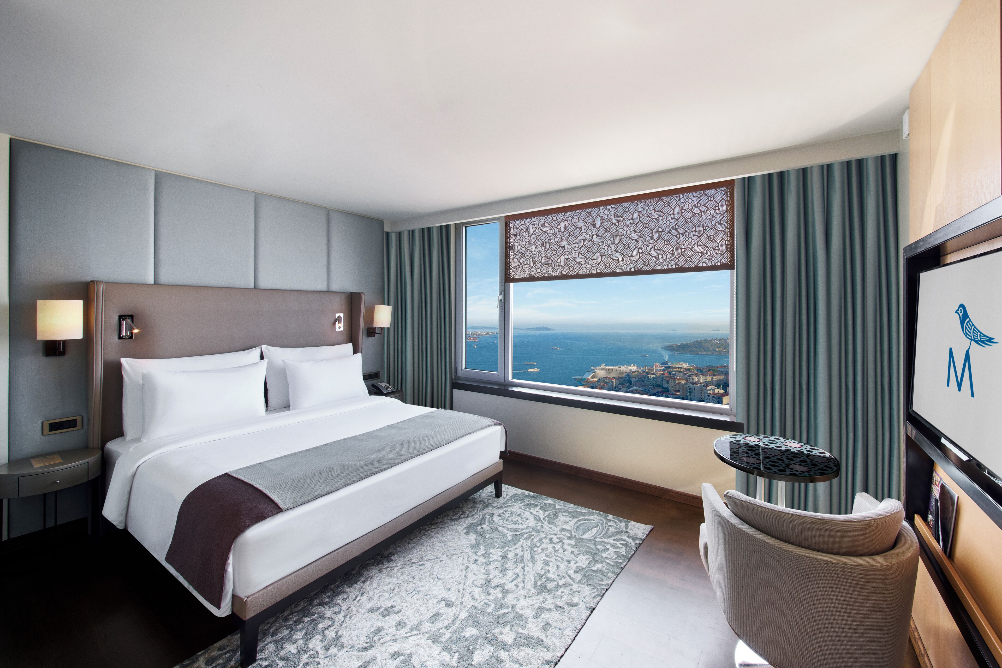 Habitación de hotel con una cama grande, vistas de la ciudad, un escritorio con lámpara y un televisor en la pared.