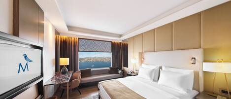 A hotel room with a bed, a desk with a chair, a TV, a window with a view, and a nightstand with a lamp.