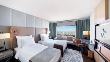 A hotel room with two beds, a desk, a chair, a TV, and a view of the city.