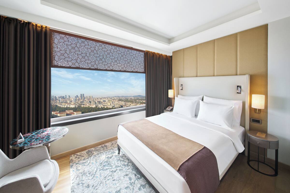 A hotel room with a large bed, a small table, a chair, and a view of a cityscape.