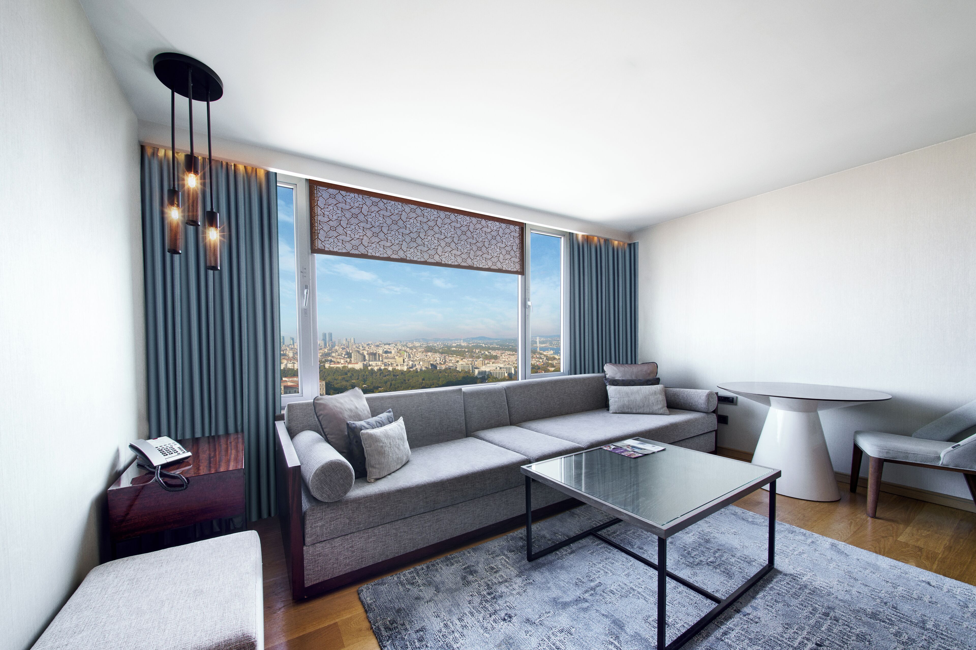 A modern living room with a grey sofa, a glass coffee table, and a view of a cityscape through large windows.