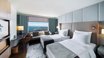 Habitación de hotel con dos camas, un escritorio con silla, televisión y un amplio ventanal con vista a la ciudad.