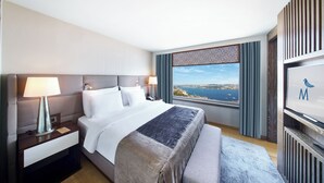 Habitación de hotel con una cama grande, una mesita de noche, una lámpara, vistas al mar y un televisor en la pared.
