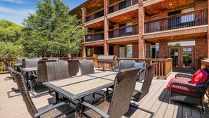 An outdoor deck with a dining area, wicker furniture, and a view of a multi-story building with brickwork and balconies.