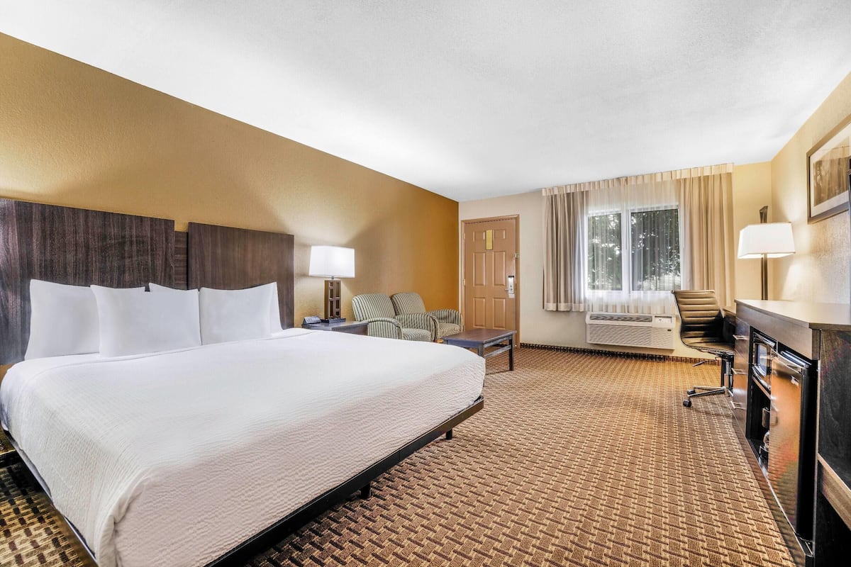 A hotel room with a large bed, a desk, a chair, a sofa, a window with curtains, and a wall-mounted air conditioning unit.