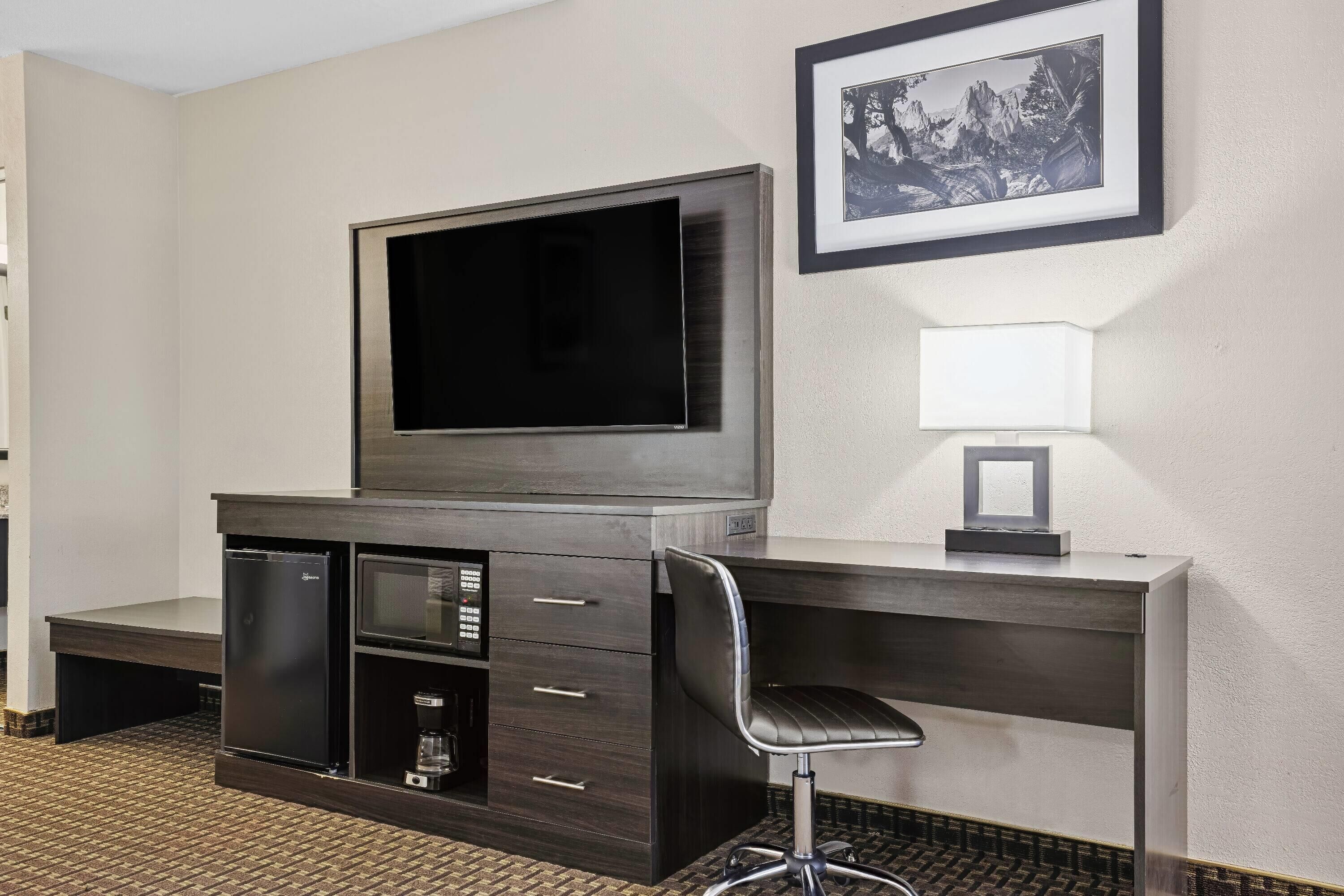 Une chambre d’hôtel avec un bureau en bois foncé, un téléviseur à écran plat, un petit réfrigérateur, une chaise, une table de chevet et un tableau encadré au mur.