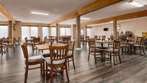 A spacious dining area with wooden tables and chairs, large windows, and a kitchen area in the background.