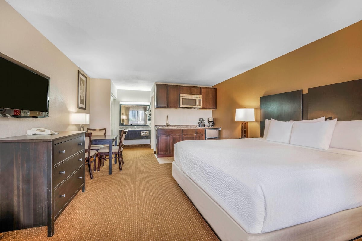 A hotel room with a large bed, a desk with a chair, a kitchenette, and a television.