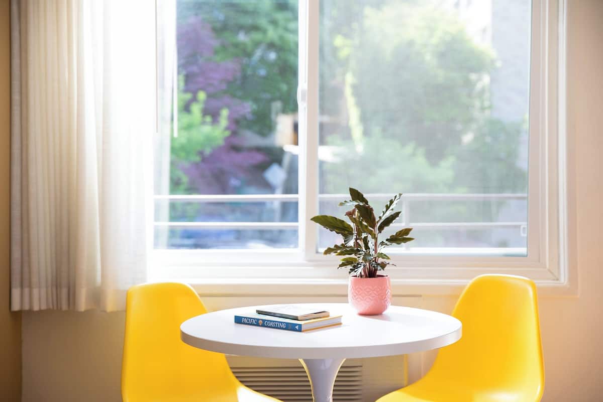 A room with a white table, two yellow chairs, a window with a view of greenery, and a pink plant on the table.