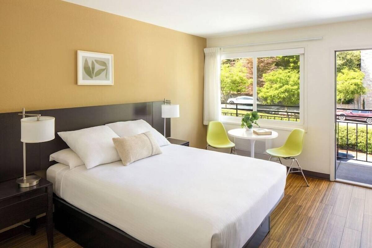A hotel room with a bed, bedside lamps, a small table with chairs, a window with a view of trees and a car, and a balcony.