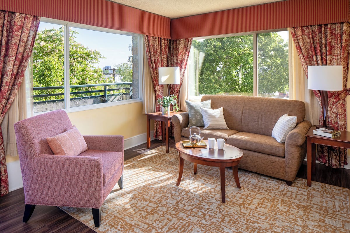 A cozy living room with a sofa, armchair, and coffee table, large windows with curtains, and a view of greenery outside.