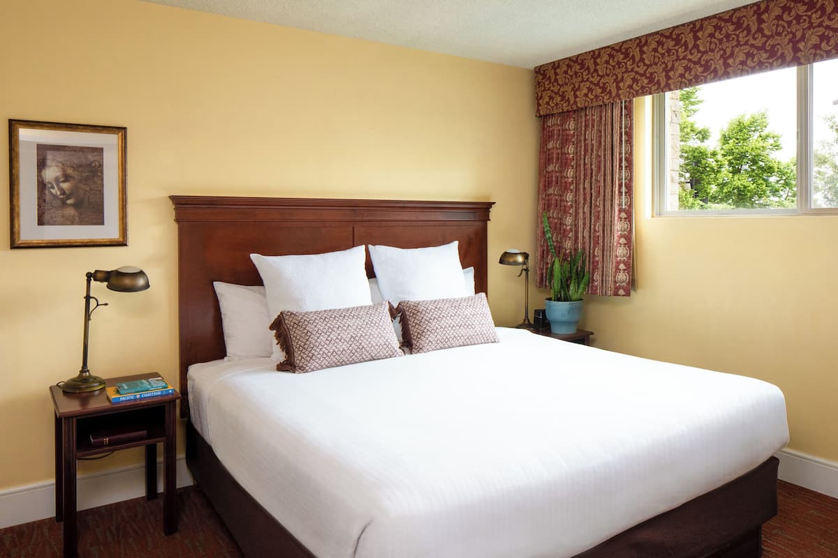 A neatly made bed with white linens and patterned pillows, a wooden headboard, a nightstand with a lamp and books, a framed picture on the wall, and a window with curtains.