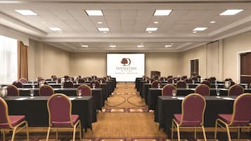 A conference room with rows of tables and chairs, a projector screen displaying a logo, and a carpeted floor.