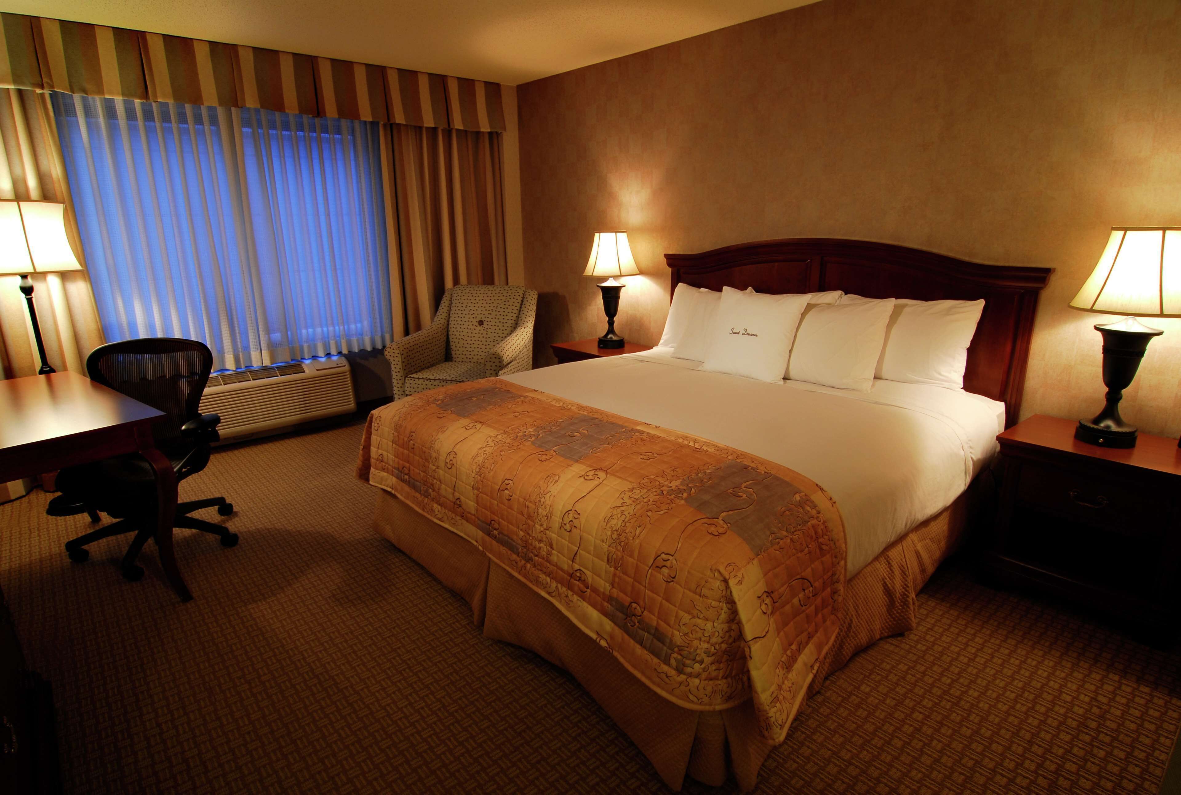 A hotel room with a large bed, two bedside lamps, a desk with a chair, and a window with curtains.