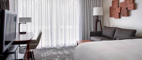 A hotel room with a large bed, a desk with a TV, a chair, a lamp, and a view of the outdoors through sheer curtains.