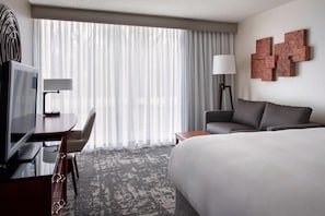 Une chambre d’hôtel avec un grand lit, un bureau avec une télévision, une chaise, une lampe et une vue sur l’extérieur à travers des rideaux transparents.