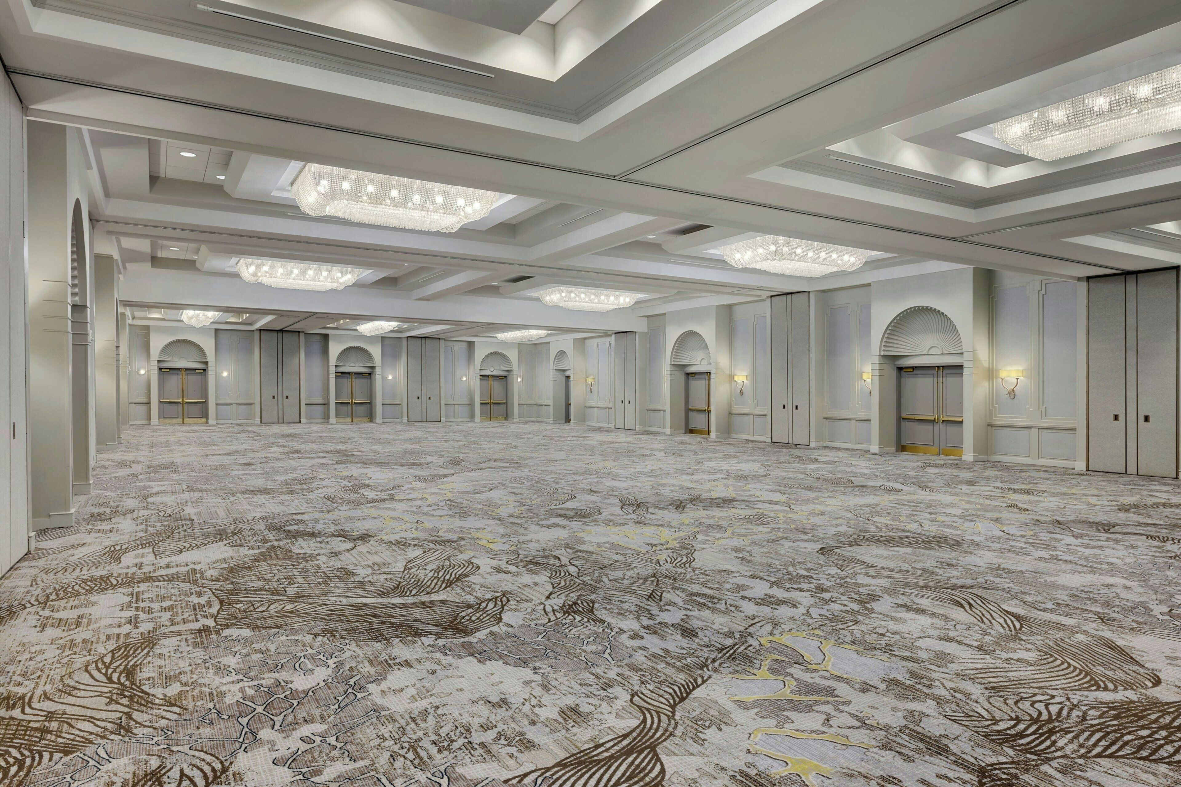 A spacious hotel lobby with a patterned carpet, multiple doors, and elegant chandeliers.