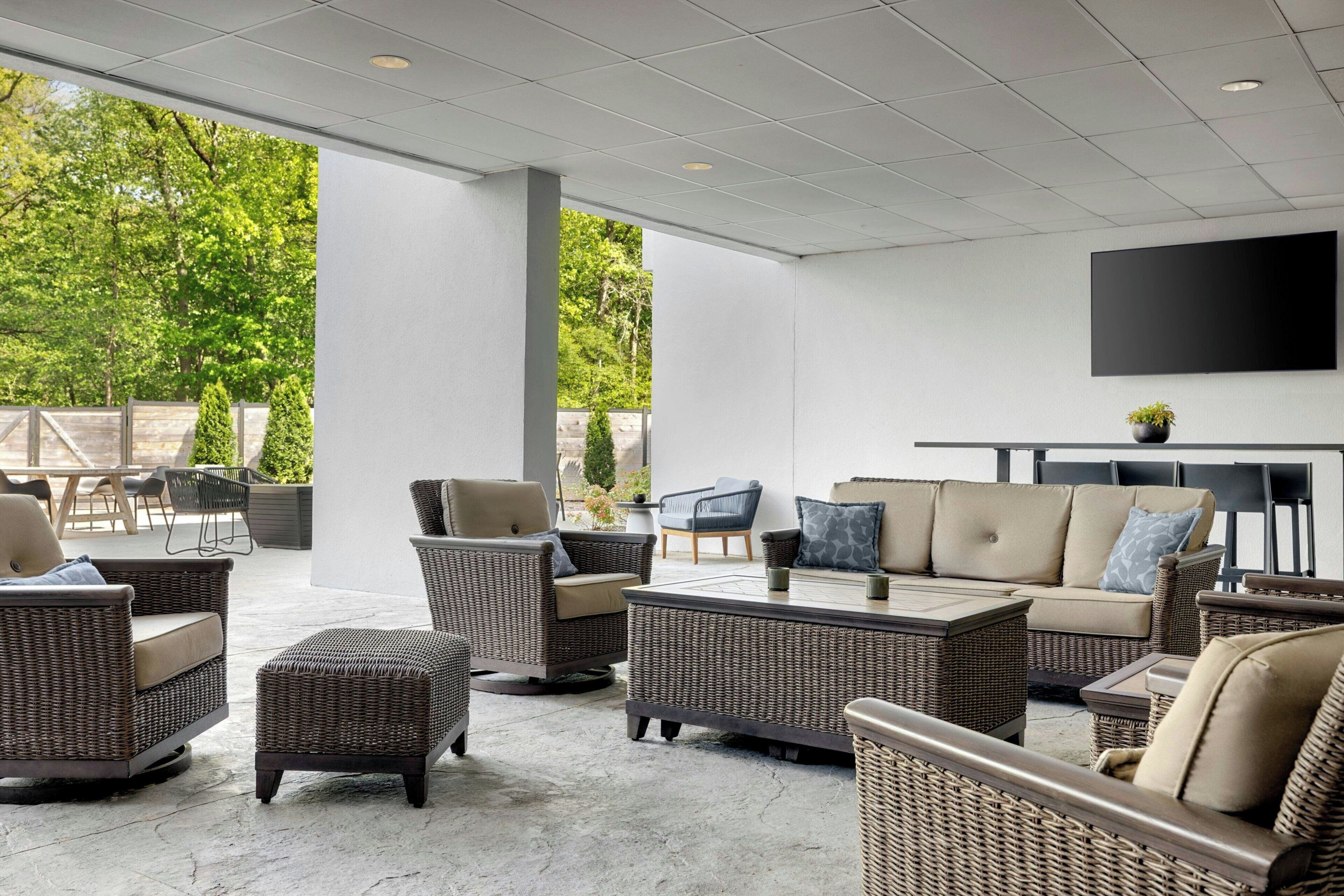 An outdoor patio area with wicker furniture, a flat-screen TV mounted on the wall, and a view of greenery.