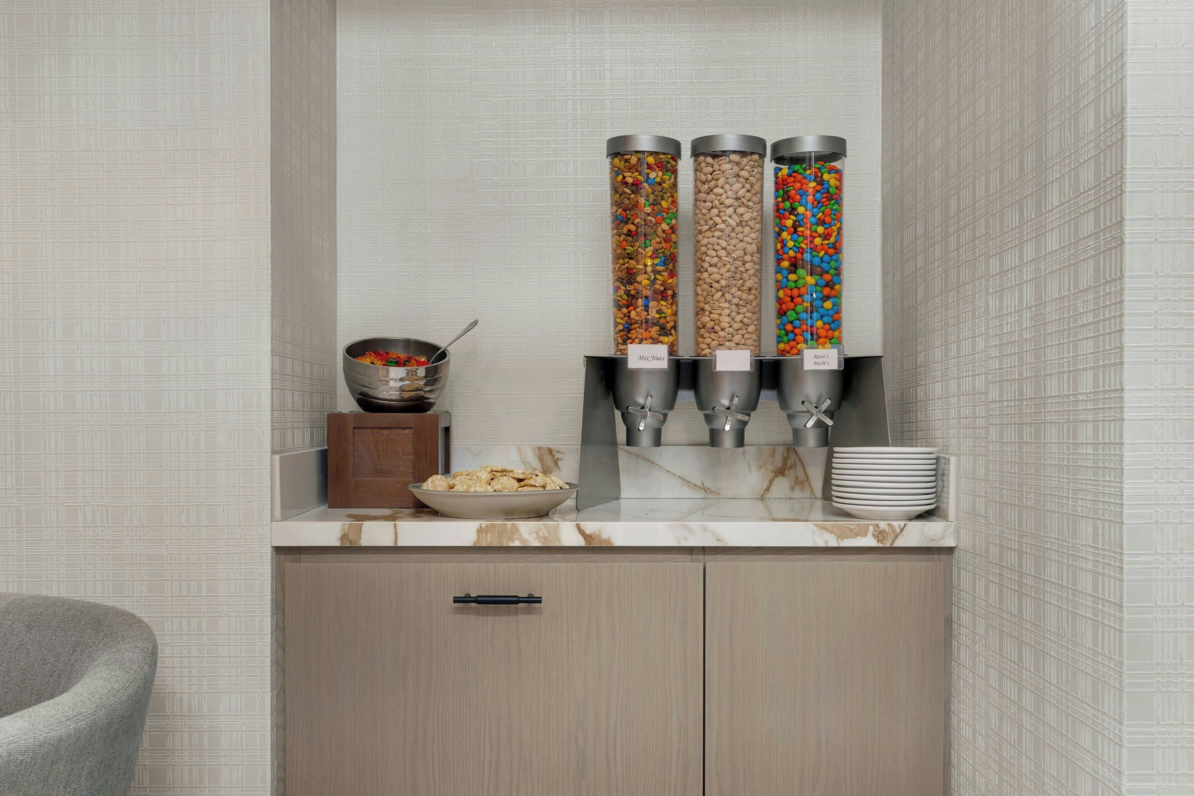 Una encimera de cocina con tapa de mármol, un mueble de madera debajo y tres dispensadores etiquetados como 'Mix Nuts' y 'M&M's'.