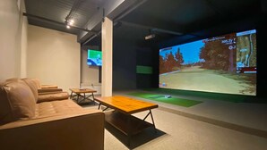 A room with a large screen displaying a golf course simulation, a brown leather sofa, a wooden coffee table, and a small table with a chair. - Ramada by Wyndham Lethbridge (Lethbridge)