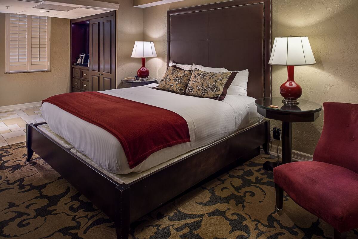 A neatly made bed with a red blanket, two bedside lamps, a red chair, and a nightstand with a phone and a lamp.