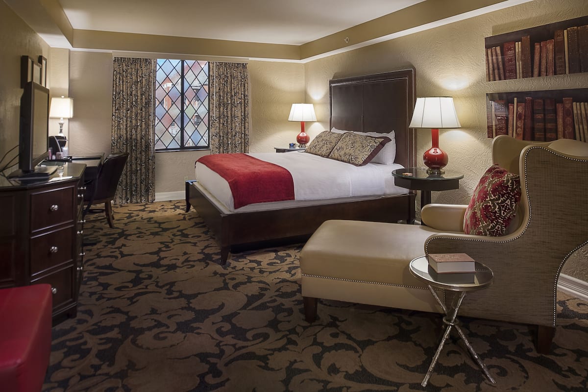 A hotel room with a large bed, a desk, a chair, a small table, and a bookshelf.
